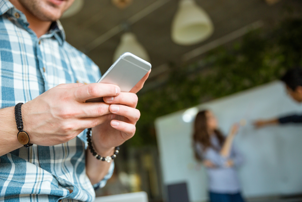 Closeup of hands of young man in checkered shirt using mobile phone while his partners arguing Closeup of hands of young man in checkered shirt using mobile phone while his partners arguing